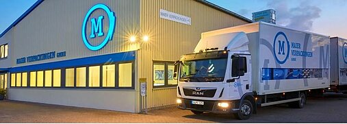 Ein LKW steht vor einer Halle bei Nacht. Auf der Halle und auf dem LKW steht das Logo und der Name von Maier Verpackungen.